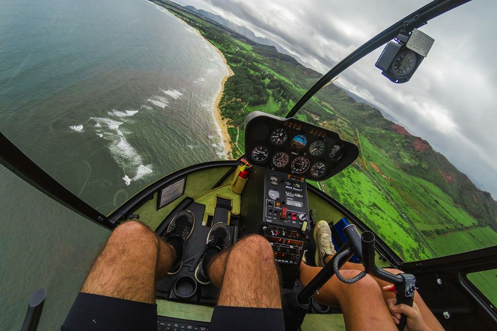 Vista de dentro da cabine de um helicóptero em voo sobre uma costa verde, com os pedais e os instrumentos em primeiro plano