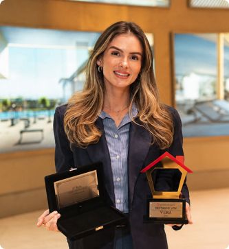 Vera Teixeira segurando um troféu de vendas, de blazer escuro, diante de uma estante clara