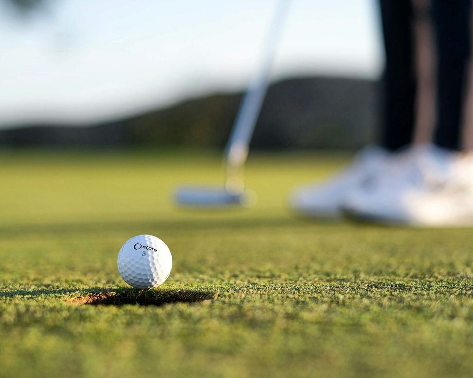 Bola de golfe sobre o green, com o taco e os pés do jogador desfocados atrás
