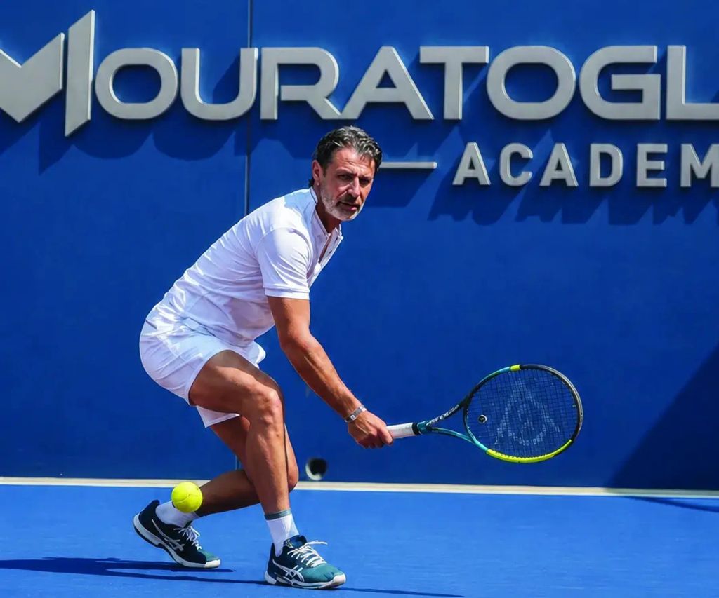 Jogador de tênis de branco em quadra rápida azul, diante do letreiro Mouratoglou Academy