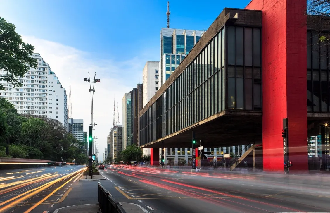 Edificio suspendido sobre pilares rojos junto a una avenida, con estelas de luz de los coches y edificios altos al fondo
