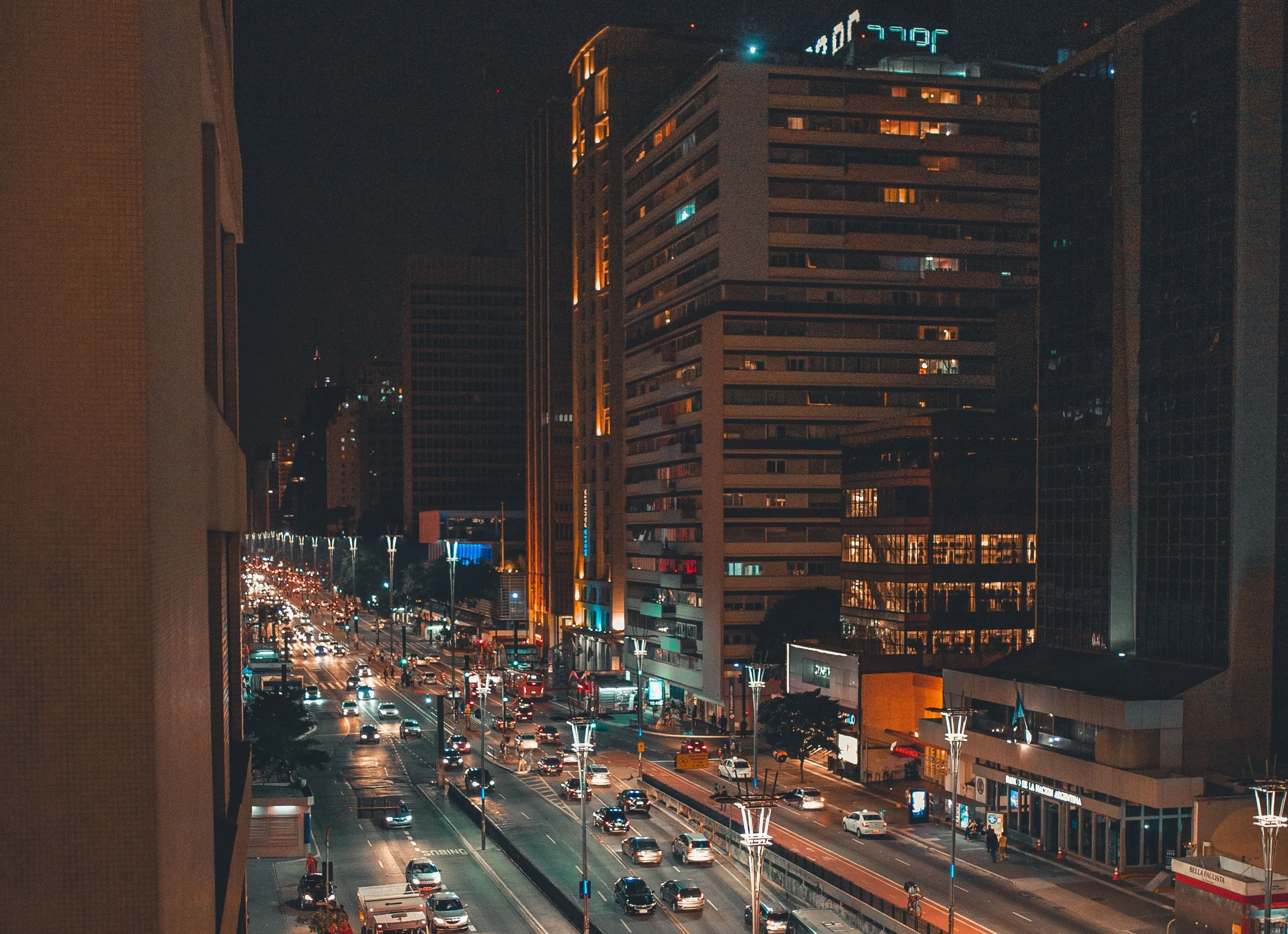 Vista nocturna, desde lo alto, de una avenida ancha con tráfico, farolas encendidas y edificios altos con ventanas iluminadas